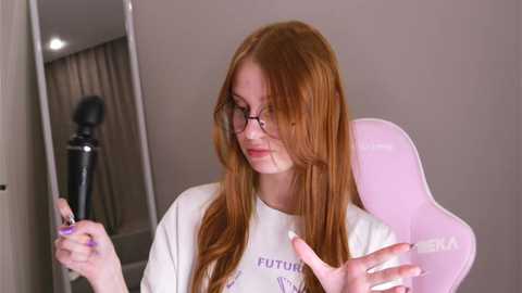 Media: Video of a red-haired woman with glasses in a white T-shirt, holding a phone in a beige room with a large mirror and a standing lamp.