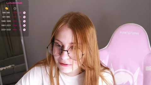 Media: Video of a young woman with long, straight, light red hair, fair skin, wearing round glasses, a white t-shirt, and a pink gaming chair.