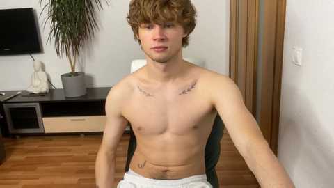 Media: Video of a shirtless, slim, young white man with curly brown hair, wearing white shorts, sitting on a chair in a modern living room with wooden floors, a black TV, and a potted plant.