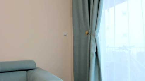 Media: A video of a minimalist room featuring a light blue sofa against a cream-colored wall. A sheer white curtain with a blue tie-back hangs to the right, partially covering a window. The room is well-lit, giving a clean and serene atmosphere.