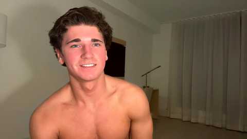 Media: Video of a shirtless, fit young man with curly brown hair, smiling, in a dimly lit room with white walls and sheer curtains.