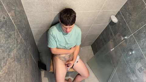 Media: A video of a young man with short dark hair, wearing a light blue T-shirt, sitting on a wooden stool in a small, tiled shower. He is using a white smartphone.