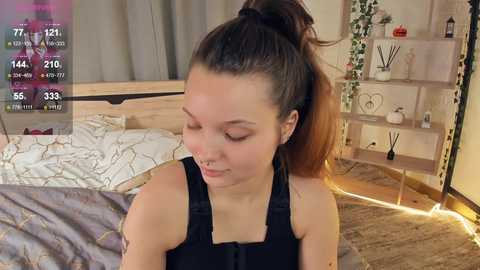 Media: Video of a young woman with fair skin and brown hair in a high ponytail, wearing a black tank top, sitting on a bed with a wooden headboard and white bedding, surrounded by a cozy, modern bedroom with wooden floor, green curtains, and a small shelf with decorative items.