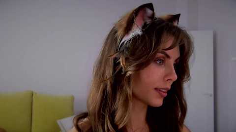 Media: Video of a fair-skinned woman with long, wavy brown hair, wearing cat ears with feathers, and a green shirt, against a plain white wall background.