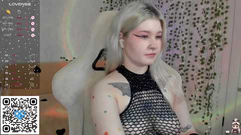 Media: Video of a young woman with platinum blonde hair, wearing a black fishnet top, sitting in a white gaming chair, surrounded by a colorful, abstract background.