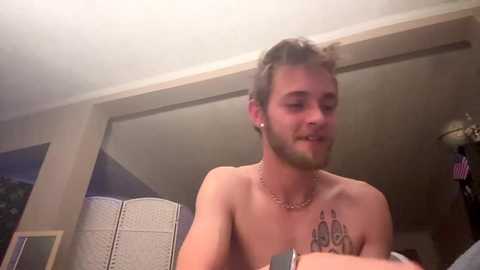 Media: A video of a shirtless man with a tattoo on his chest, short brown hair, and a beard, smiling in a bedroom with a mirror, white walls, and a bed in the background.