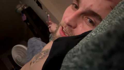 Media: Video of a young man with light skin and short brown hair, wearing a black t-shirt, lying on a bed with a grey blanket. His face is close to the camera, smiling slightly.