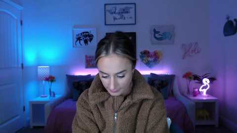 Media: A video of a young woman in a brown hoodie, standing in a dimly lit, purple-lit bedroom with a bed, two side tables, and decorative items.