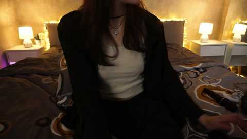 Media: Video of a young woman with long, dark hair, wearing a black cardigan over a white top, sitting on a bed with patterned sheets. Warm, ambient lighting from bedside lamps creates a cozy atmosphere.