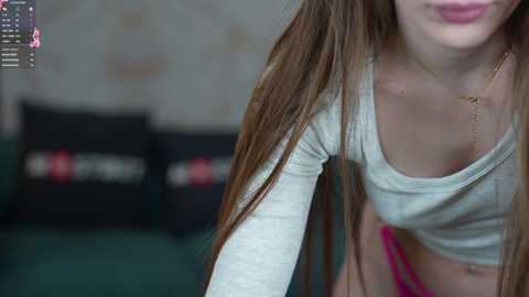 Media: Video of a young woman with long brown hair, wearing a light grey crop top and pink panties, leaning forward in a casual, slightly blurry room with dark pillows.