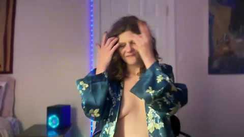 Media: Video of a topless woman with long brown hair, wearing an unbuttoned floral robe, covering her face with both hands, in a dimly lit room with a blue neon strip light, framed artwork, and a speaker.