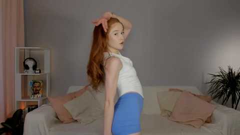 Media: Video of a slender, fair-skinned woman with long red hair, wearing a white tank top and blue shorts, standing in a minimalist living room with beige walls, a white couch, and a small black plant.