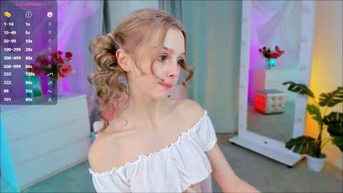 Media: Video of a young Caucasian woman with fair skin and blonde curls in pigtails, wearing a white off-shoulder top, indoors with colorful lighting, a potted plant, and a mirror.