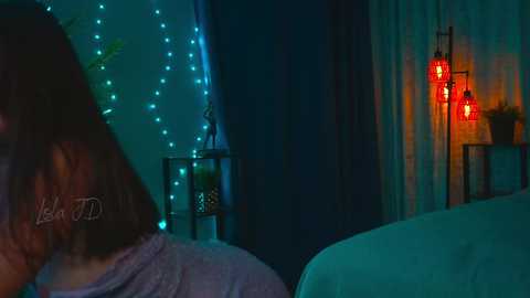 Media: A video of a dimly-lit room with a teal light, featuring a person with shoulder-length brown hair, a blue shirt, and a visible tattoo, sitting in a chair. Red lanterns and string lights add a cozy ambiance.