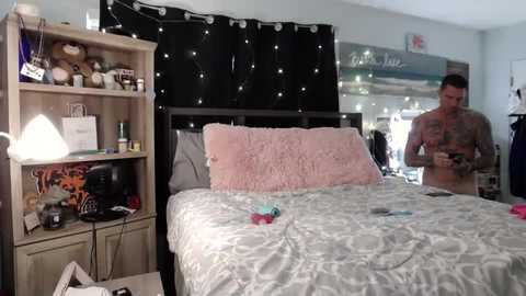 Media: Video of a messy bedroom with a tattooed man in the background, a plush pink pillow, a gaming console, and a black curtain with fairy lights.