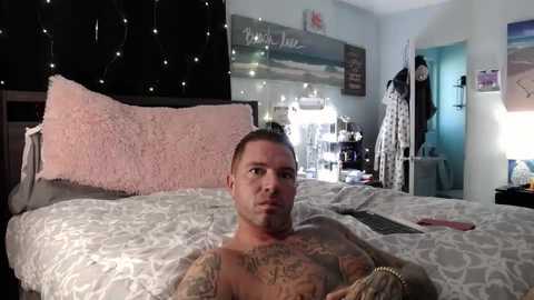 Media: Video of a shirtless, tattooed man with a serious expression lying on a bed with white, pink, and gray bedding. The room has a cozy, slightly messy atmosphere with fairy lights, a computer, and a hanging jacket.