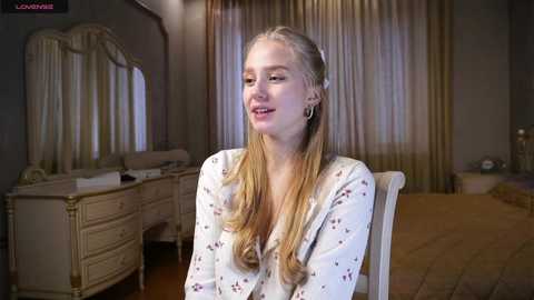 Media: Video of a smiling blonde woman with long hair, wearing a white floral robe, seated in a luxurious bedroom with cream curtains, a dresser, and bed.