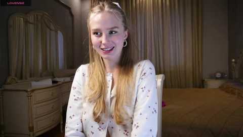 Media: Video of a smiling, blonde-haired young woman with fair skin, wearing a white floral blouse, seated in a luxurious, dimly lit bedroom with beige curtains and a golden bedspread.