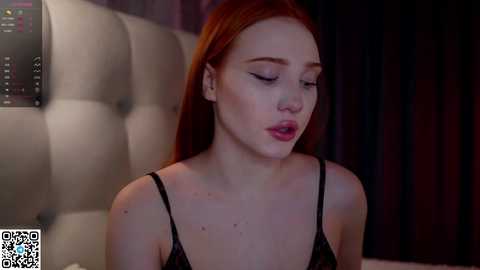 Media: Video of a fair-skinned, red-haired woman with closed eyes, wearing a black lace camisole, in a dimly lit room with a white, tufted headboard.