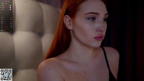 Media: Video of a pale-skinned, red-haired young woman with long hair, wearing a black strapless top, gazing contemplatively. Background features a dimly lit, tufted leather headboard.