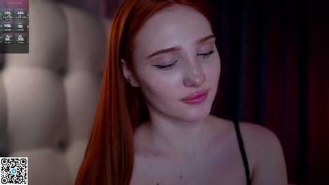 Media: A video of a fair-skinned woman with long, straight red hair, wearing a black spaghetti strap top, softly smiling and closing her eyes. The background features a dimly lit, plush, tufted headboard.