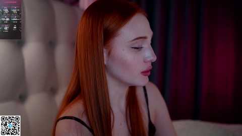 Media: Video of a pale-skinned, red-haired woman with long, straight hair, wearing a black bra, in a dimly lit room with a plush, tufted white headboard and dark curtains.