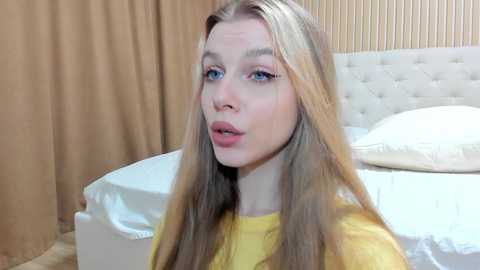 Media: A video of a young woman with long, straight blonde hair, fair skin, and blue eyes, wearing a yellow shirt, sitting on a bed with white sheets and a beige headboard in a softly lit bedroom.