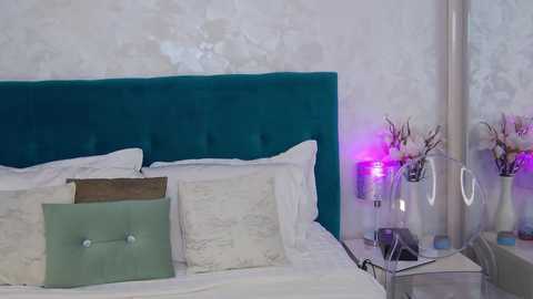 Media: Video of a modern bedroom featuring a teal velvet headboard, white bedding, a green pillow, floral-patterned wallpaper, a glass bedside table with purple flowers, and a silver lamp.