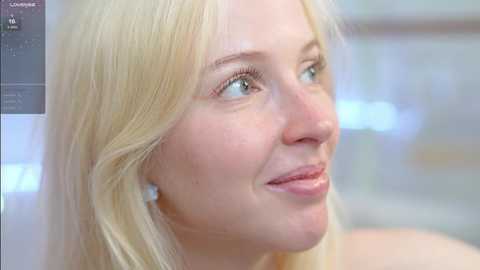 Media: Video of a fair-skinned, blonde woman with light green eyes, smiling softly. She wears subtle makeup and has a soft, natural look. The background is blurred, suggesting an indoor setting with soft lighting.