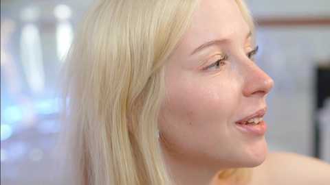 Media: Video of a smiling Caucasian woman with light skin, straight blonde hair, and fair complexion. She is looking to the right, in a blurred indoor setting with white walls and a hint of greenery.