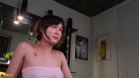 Media: Video of a young woman with fair skin and short dark hair, wearing a strapless pink top, in a modern bathroom with a mirror, plants, and abstract art on the wall.