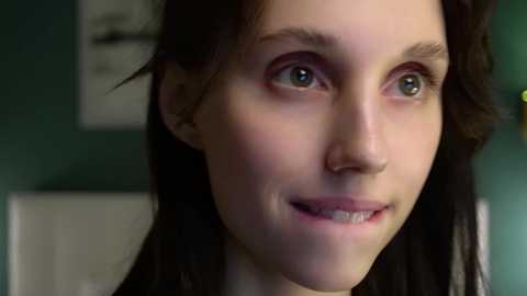 Media: Video of a young woman with fair skin and brown eyes, smiling, with straight dark hair, in a teal room with a white wall and a blurred white object in the background.