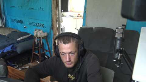 Media: Video of a young man with short hair wearing a black hoodie, headphones, and a microphone, sitting in a cluttered, blue-walled room with a bed, shelves, and a desk.