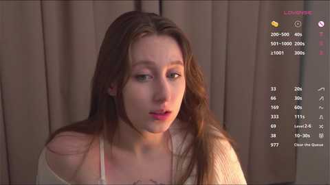 Media: Video of a young Caucasian woman with long brown hair, wearing a white off-shoulder top, looking slightly confused, against a beige curtain background.