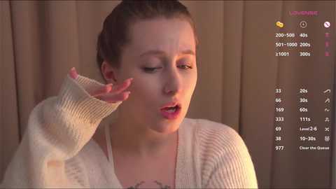 Media: Video of a fair-skinned woman with light brown hair in a bun, wearing a cream sweater, smiling and covering her mouth, against a beige curtain backdrop.