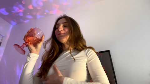 Media: A video of a woman with long brown hair, wearing a white long-sleeve top, holding a pink glass, illuminated by colorful disco lights in a dimly lit room.