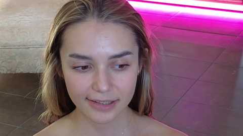 Media: Video of a young Caucasian woman with fair skin and straight, shoulder-length blonde hair. She has a natural, neutral expression. The background features a modern tiled floor and neon pink and white lighting.