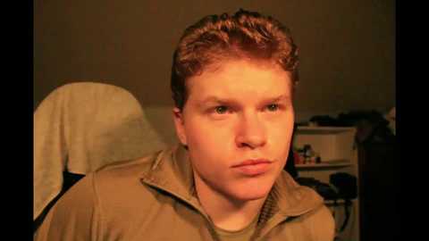 Media: Video of a young man with curly red hair, wearing a beige jacket, sitting in a beige chair against a dimly lit background.