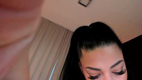 Media: Video of a woman with long, straight black hair in a high ponytail, wearing dramatic eye makeup and a low-cut top, partially visible. Background includes beige curtains and a ceiling light.