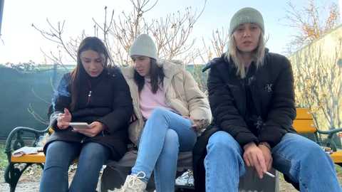 Media: Video of three young women in winter coats and hats, sitting on a bench outdoors, focused on smartphones, with a leafless tree and green fence background.