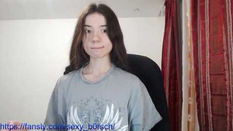 Media: A young woman with long brown hair, fair skin, and a slender physique, wearing a gray T-shirt with a graphic design, sits in a black chair against a plain white wall. Red curtains are partially visible in the background.