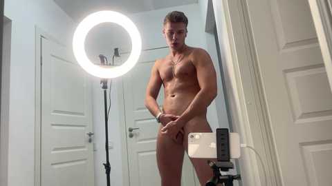 Media: Video of a muscular, light-skinned, nude man with short brown hair, holding a phone, in a brightly lit bathroom with a ring light, white door, and white walls.