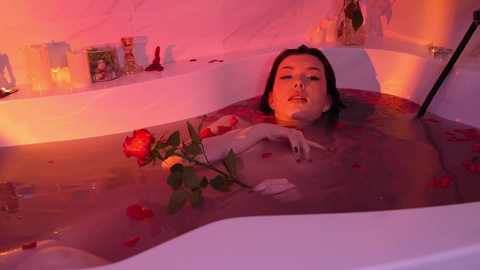 Media: Video of a woman with wet, dark hair submerged in a pink-tinted bathtub, surrounded by rose petals and candles.