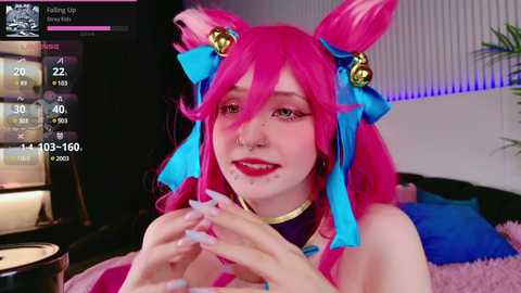 Media: Video of a young woman with pink hair, wearing a blue ribbon and cat ears, playing a video game. Background includes a bed and a screen displaying game statistics.