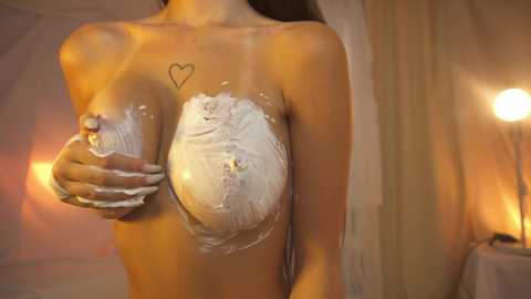 Media: A video of a topless woman with light brown skin, covered in white cream, holding her breasts. She has a small heart tattoo on her chest. Background features a softly lit, cozy room with warm colors and curtains.