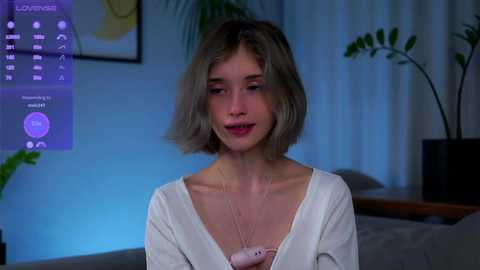 Media: Video of a young woman with short, light brown hair, wearing a white cardigan, standing in a dimly lit room with a plant and a framed picture on the wall.