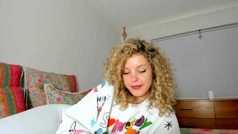 Media: Video of a curly-haired woman with fair skin and a warm smile, wrapped in a white blanket with colorful cartoon characters, in a bright, cozy bedroom with floral pillows and a wooden dresser.