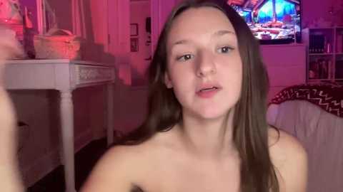 Media: Video of a young, light-skinned, topless woman with long brown hair, sitting in a dimly lit bedroom with pink lighting. TV screen and white dresser visible in background.