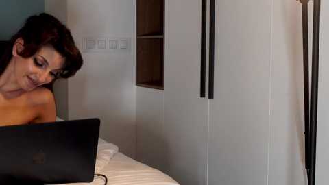 Media: A video of a woman with short brown hair, smiling, working on a laptop in a modern, minimalist bedroom with white walls, black vertical blinds, and a white bed.