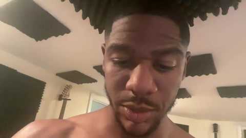 Media: Video of a shirtless Black man with a shaved head and short beard, wearing a bandage on his nose, standing indoors with a white ceiling and black acoustic panels.